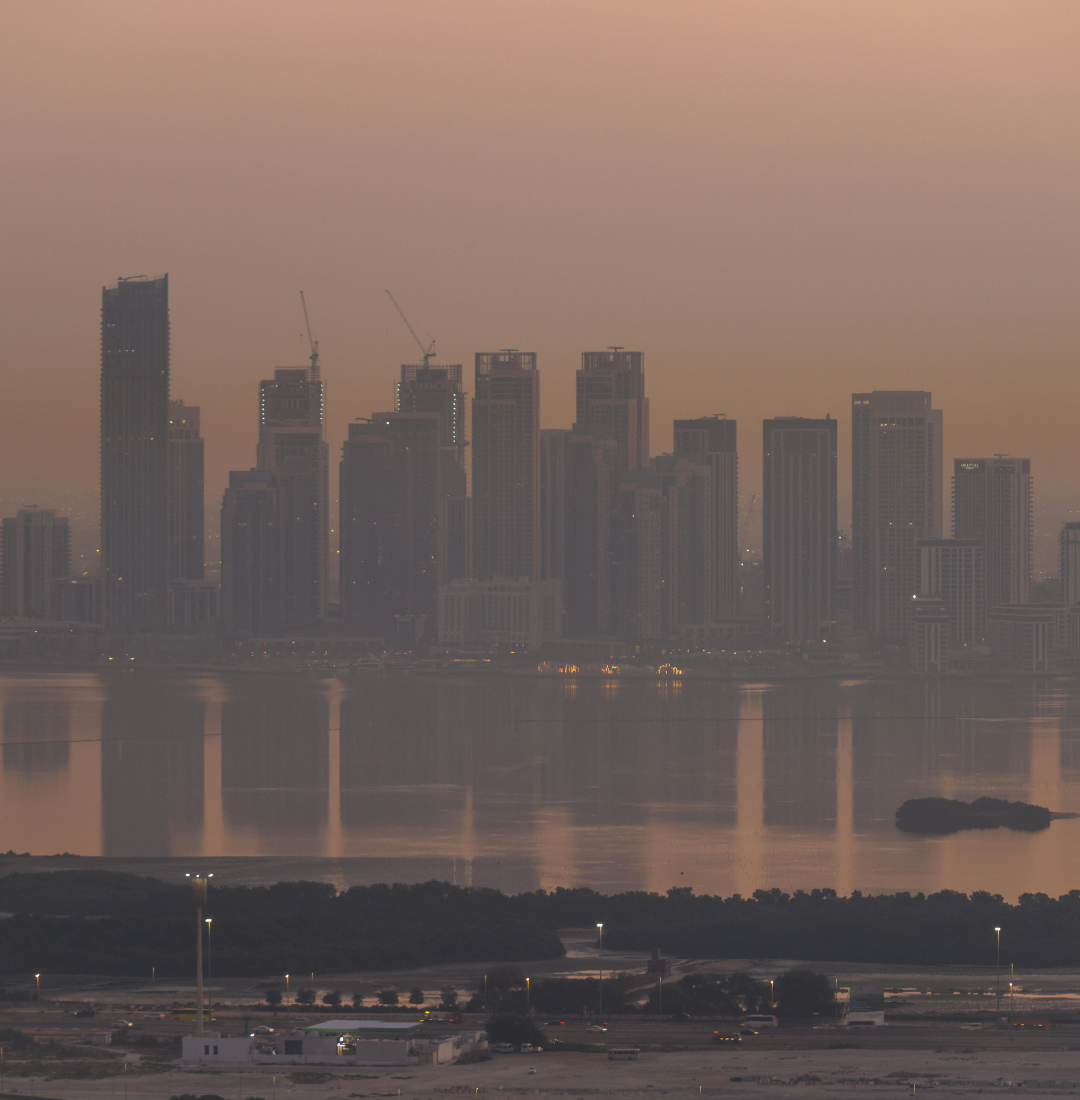 A hazy skyline of modern skyscrapers reflects in the calm water at dusk.