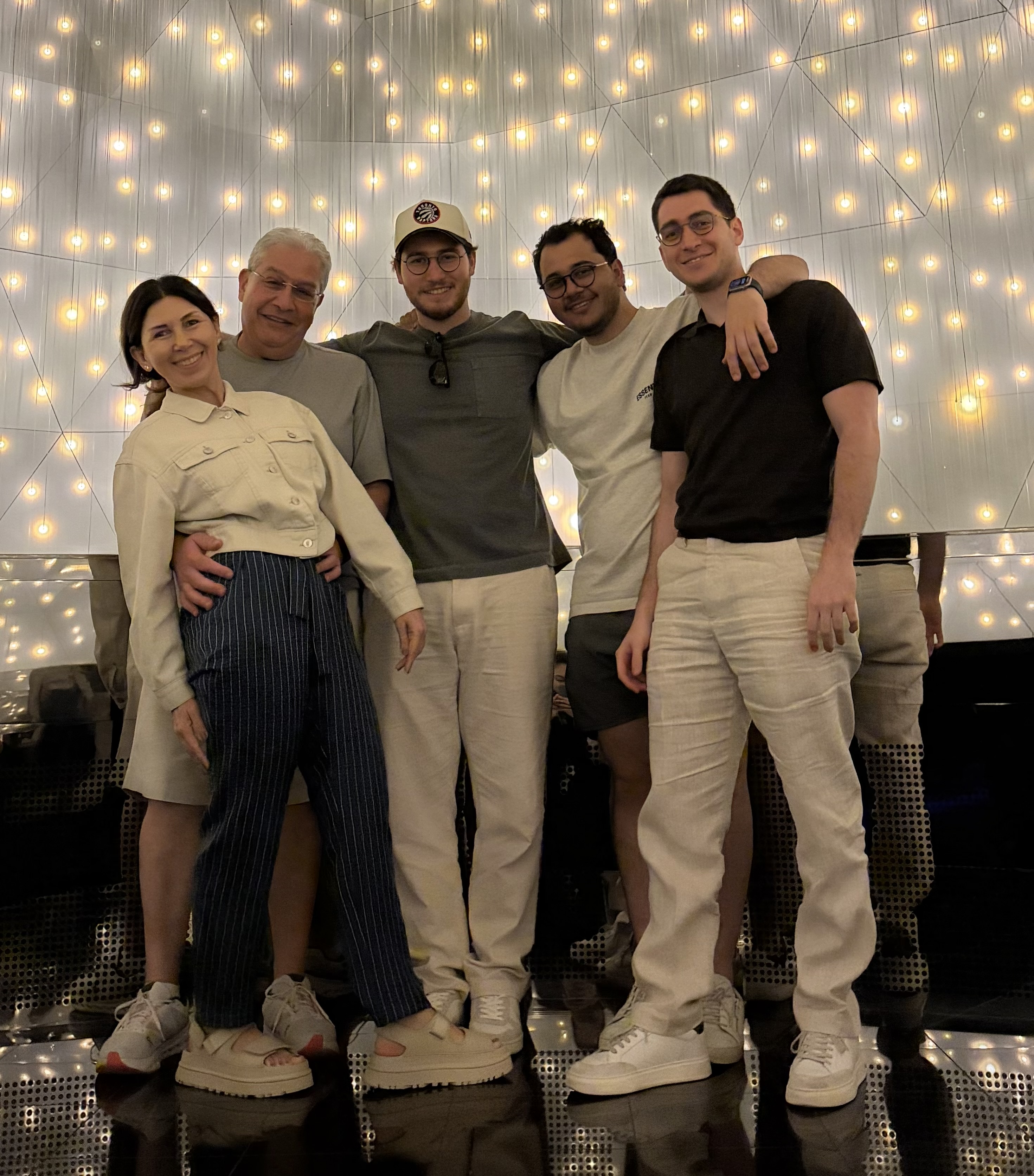 Five people stand together with their arms around each other, in front of a geometric backdrop. On the left are a man with white hair, his arms around a woman with short brown hair. On the left are 3 young men who share a strong facial resemblance and have short-cropped brown hair and glasses. 