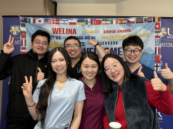 Six people of Chinese descent pose with peace signs and thumbs-up gestutes. They are standing in front of a backdrop with many flags and "Welcome" written in many languages.