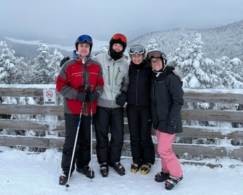 Four people bundled in winter clothing stand in front of a wooden railing overlooking snowy tree-covered mountains.