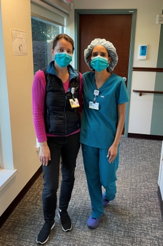 A woman wearing scrubs stands next to a woman wearing a sweater, in the hallway of a clinic.