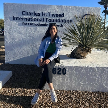 A young woman sits in front of a sign reading "Charles H. Tweed International Foundation for Orthodontic Research"