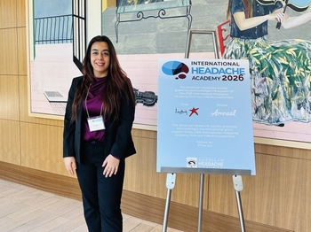 A woman wearing a pantsuit stands next to a sign reading "International Headache Academy 2026".