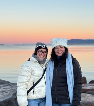 Two women with long, dark, curly hair bundled in thick winter coats and hats. They stand in front of a lake reflecting a soft orange sunset.