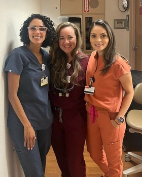 Three women wearing scrubs smile together in the clinic.