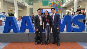 Three people in suits stand together in a large atrium, in front of a large display spelling out "AAOMS".