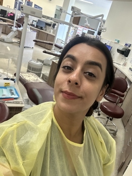 A woman wearing a yellow dental gown. Behind her is a large clinic room with multiple dental chairs arranged in a circle.