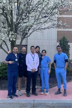 Six people stand together outside a brick building, underneath a tree with white flowers.