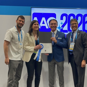 A young woman accepts a certificate and blue ribbon from a man in front of a sign reading "AAO 2026". Two other men stand smiling on either side.