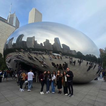 Six people pose together in front of a huge metallic sculpture shaped like a jellybean. Reflected on the surface of the sculpture and behind it can be seen many tall city buildings.