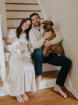 A young man and woman sit on the bottom of a staircase, leaning together. The woman holds a small baby in one arm and the man hugs a large brown dog who is sitting on his lap.