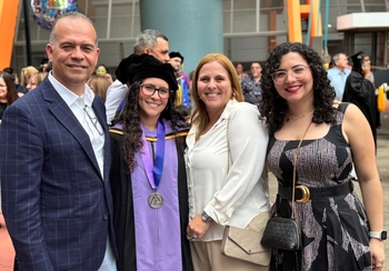 A man with short grey hair and a blonde woman pose with two young women who both have long dark curly hair. One of the young women  wears a black and purple graduation gown, a black velvet doctoral cap, and a medal on a wide purple ribbon.
