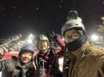Three people stand together on a snowy slope, wearing helmets and thick coats, and holding skis and ski poles.