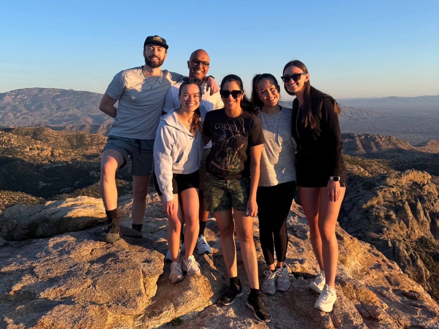 Six people stand together on an outcropping of rock. Behind them a landscape of wide plateaus stretches far into the horizon and a clear blue sky.