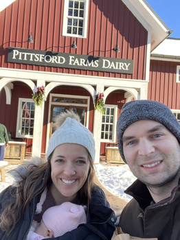 A man and a woman holding a baby stand in front of a red building, Pittsford Farms Dairy.