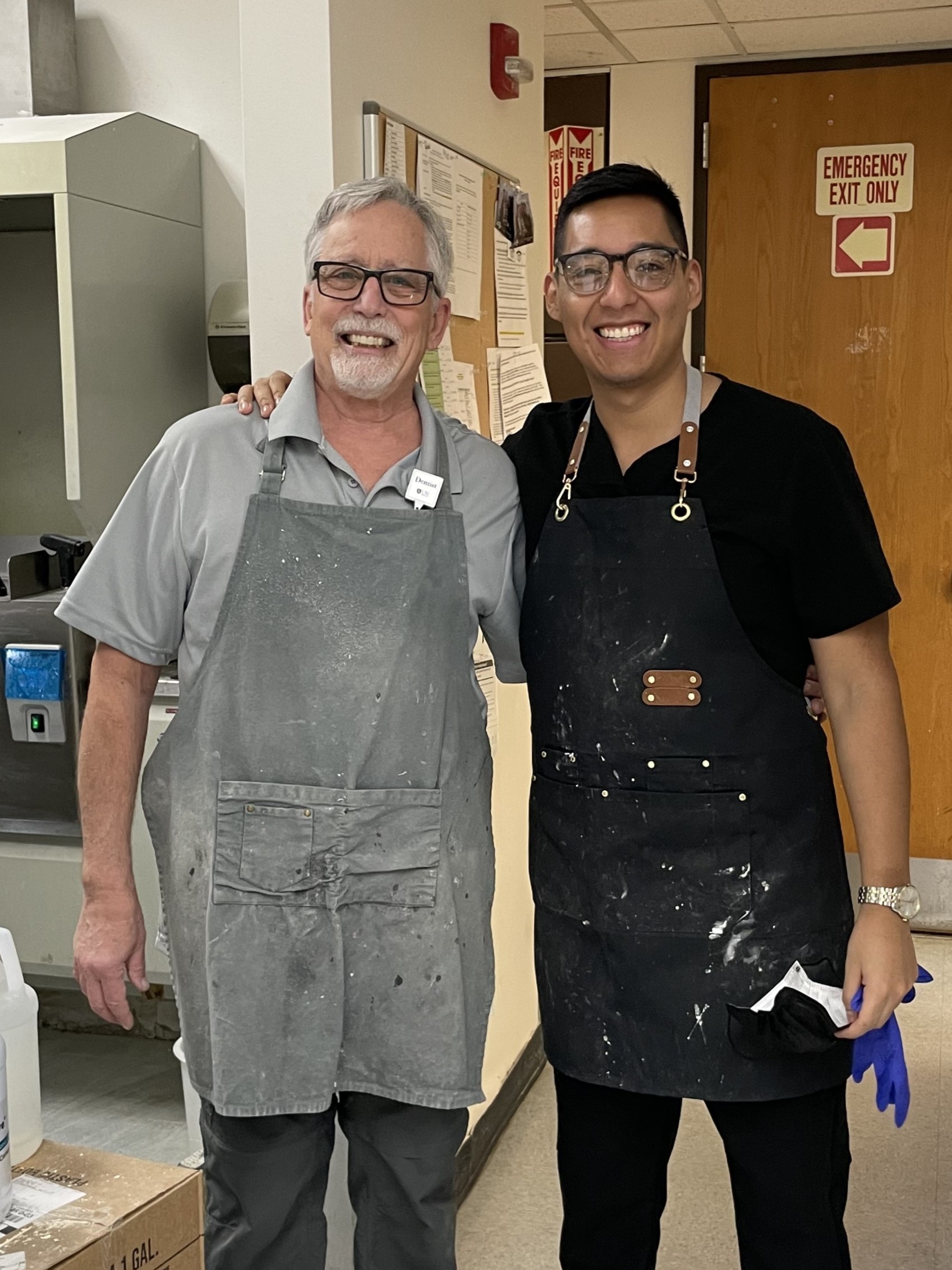 Two men stand together in a laboratory room. Both wear aprons speckled with white.