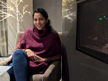 A woman with light brown skin and long brown hair smiles from an armchair next to an electric fireplace. She is dressed in jeans and a cozy maroon poncho.