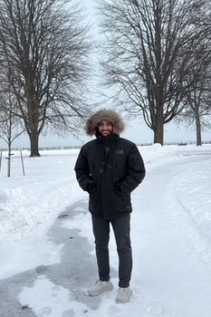 A bearded man with brown skin stands on a a snowy path, bundled in a puffy winter jacket. Behind him are a few trees bare of leaves and a pale grey sky.