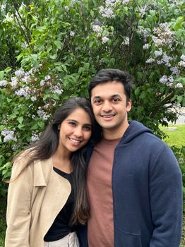 A young man and woman smile together in front of a large lilac bush with very pale flowers.