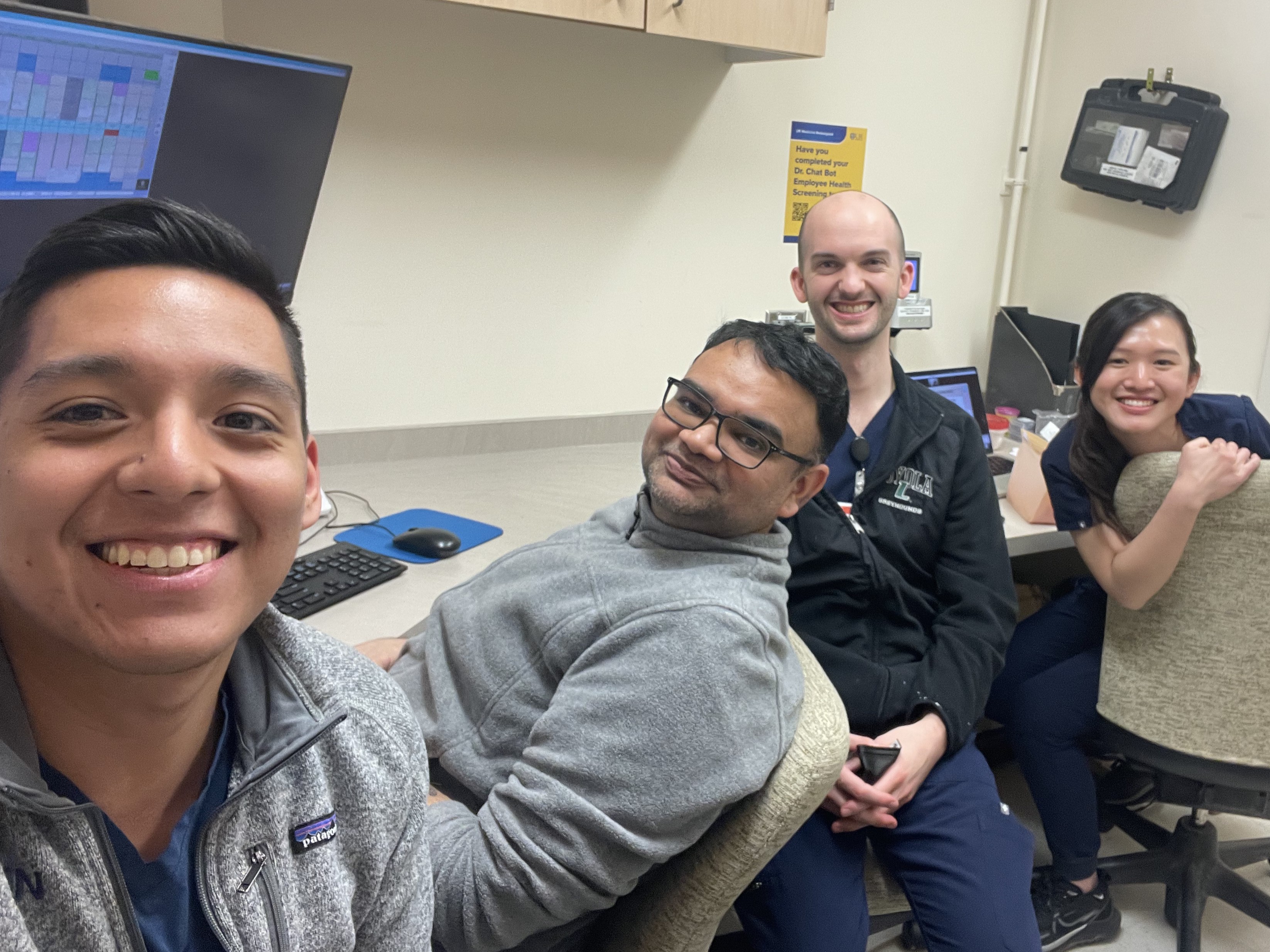 Four people sitting in front of computers on a long desk. They are all turned around toward the camera, smiling.