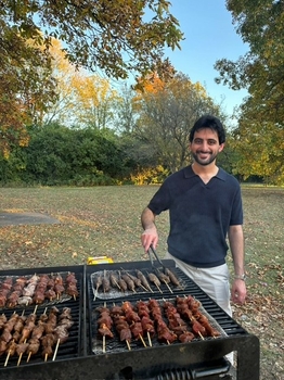 A bearded man smiles as he cooks skewers of meat on a large grill. Behind him is a small grass clearing surrounded by autumn trees.