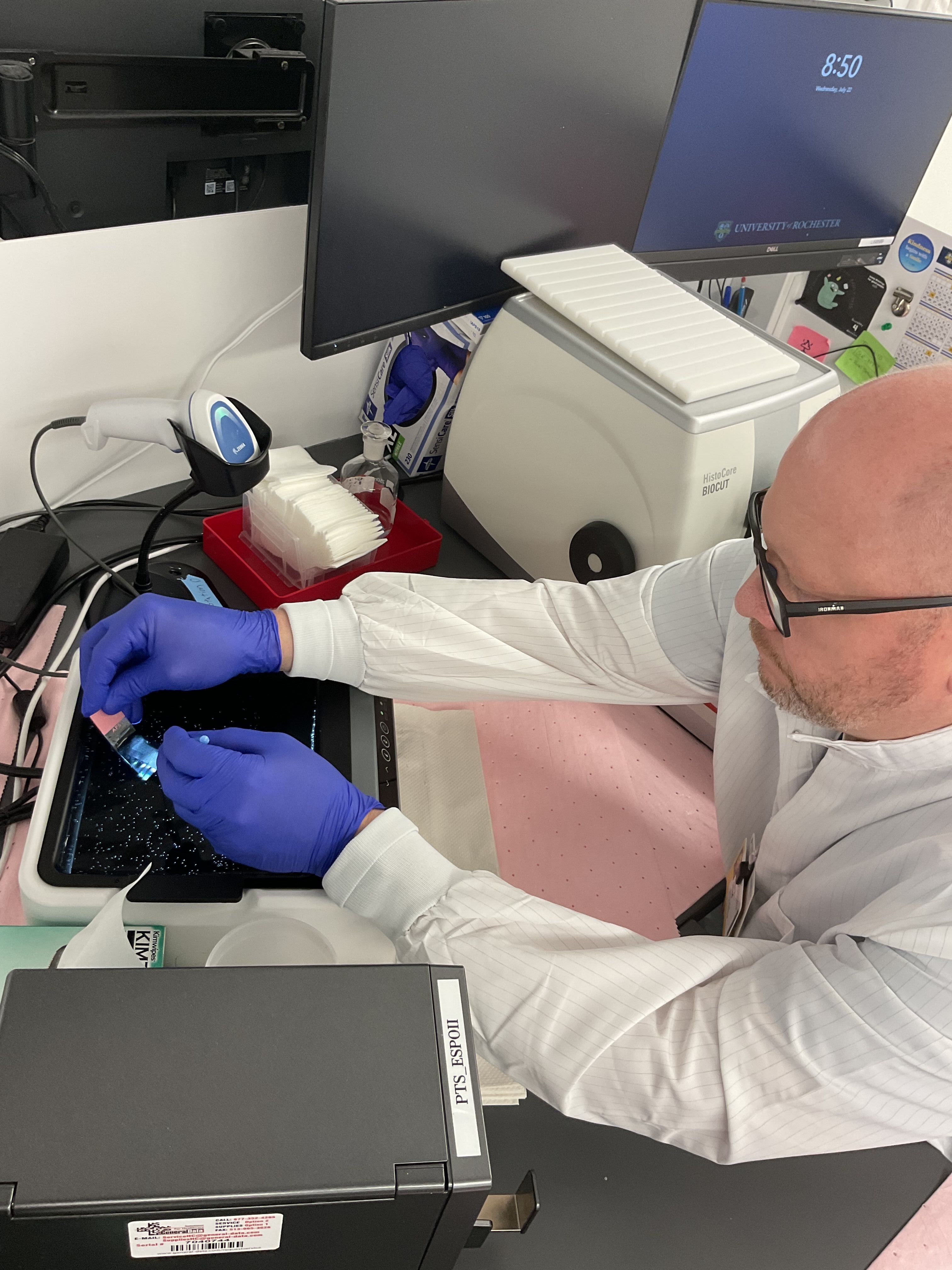 Dr. Toland in white lab coat and gloves is seated and working with a sample