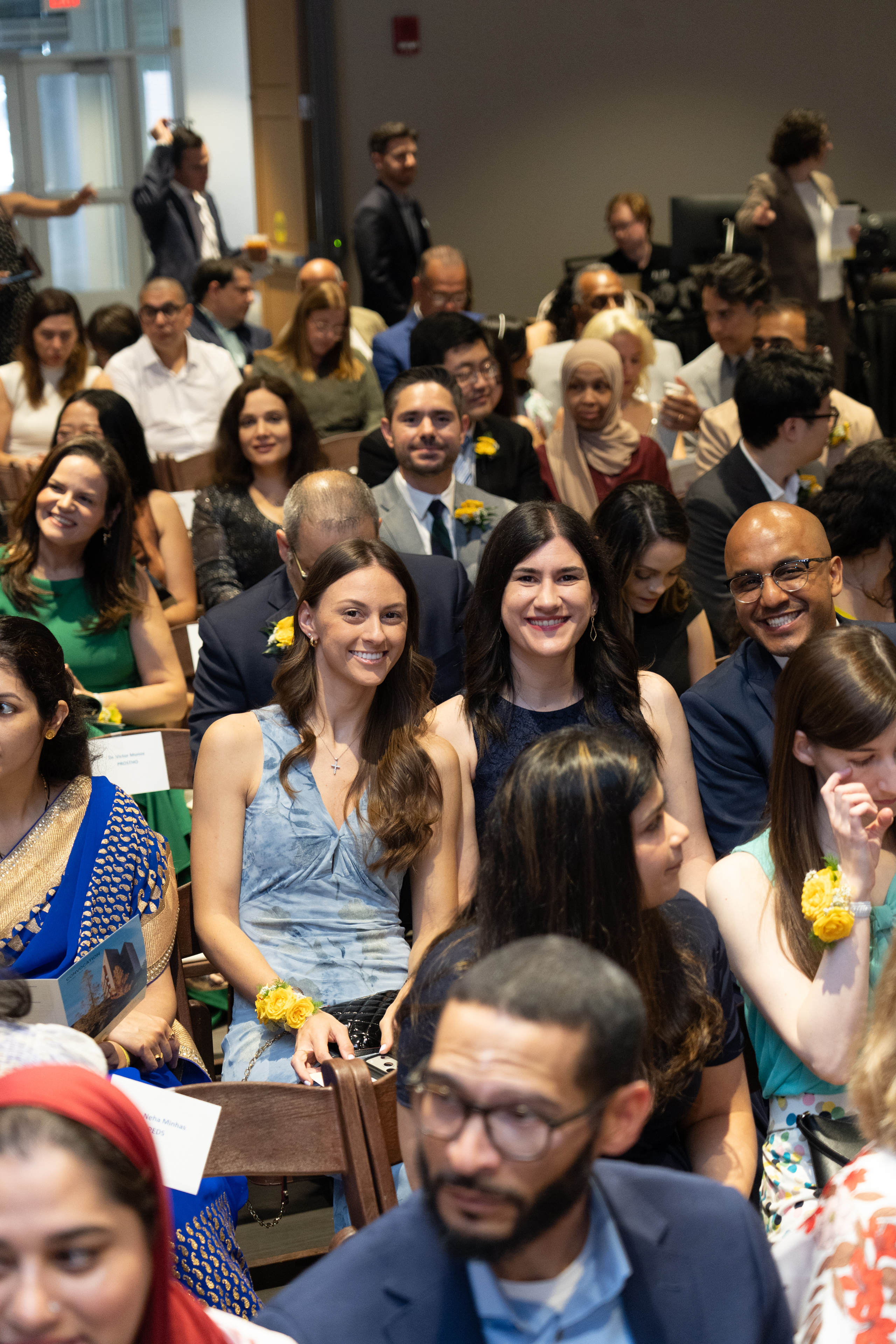 Three young people smile from the middle of a crowd. The group is quite varied in ethnicities and clothing styles.