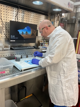 Jason Toland wearing a white lab coat, safety glasses, and blue gloves prepares a tissue specimen at a stainless steel grossing station. Using forceps, the individual carefully handles a sample beside labeled containers and a pathology cassette while working in a ventilated laboratory workspace.