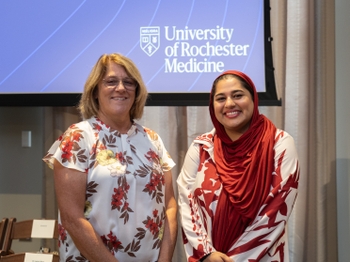 On the left, a middle-aged woman with pale skin, shoulder-length blonde hair, and glasses. On the right, a young woman with light brown skin and dark hair pulled back under a flowing red headscarf.