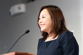 A woman of Asian descent with shoulder-length brown hair smiles in front of a podium microphone.