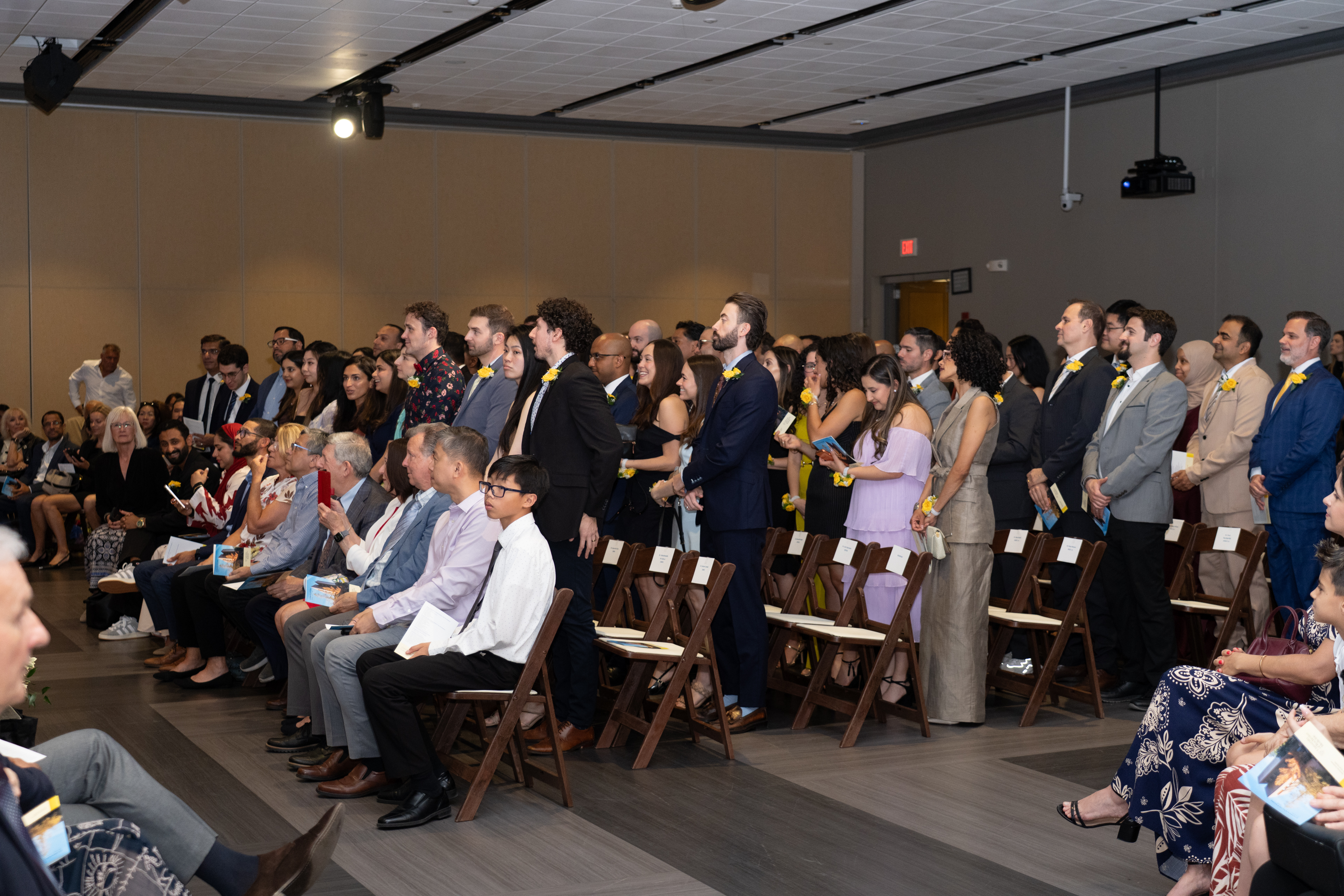 Multiple rows of people wearing yellow rose corsages stand in front of their seats in a large room.