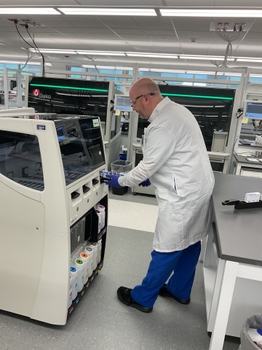 Jason Toland operating a large automated machine in a clinical or research lab setting. Technician wears white lab coat, blue gloves, and blue pants, with multiple machines and computer monitors visible in a clean, well-lit environment.