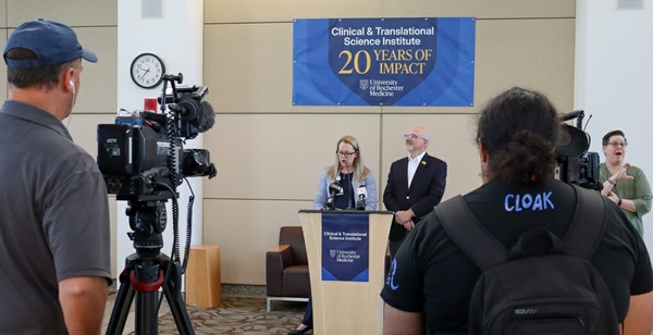 Karen Wilson and Martin Zand standing at a podium with a celebration banner above them that reads 20 Years of Impact. Two camera persons are in the foreground.