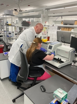 Two laboratory technicians working with a scientific instrument, likely a microscope or analyzer, in a clinical or research lab setting. The scene includes lab equipment, a computer mouse, and various supplies on a gray countertop, highlighting collaboration and technical analysis.