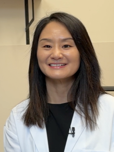 A smiling woman of Asian descent with dark hair, wearing a lab coat.