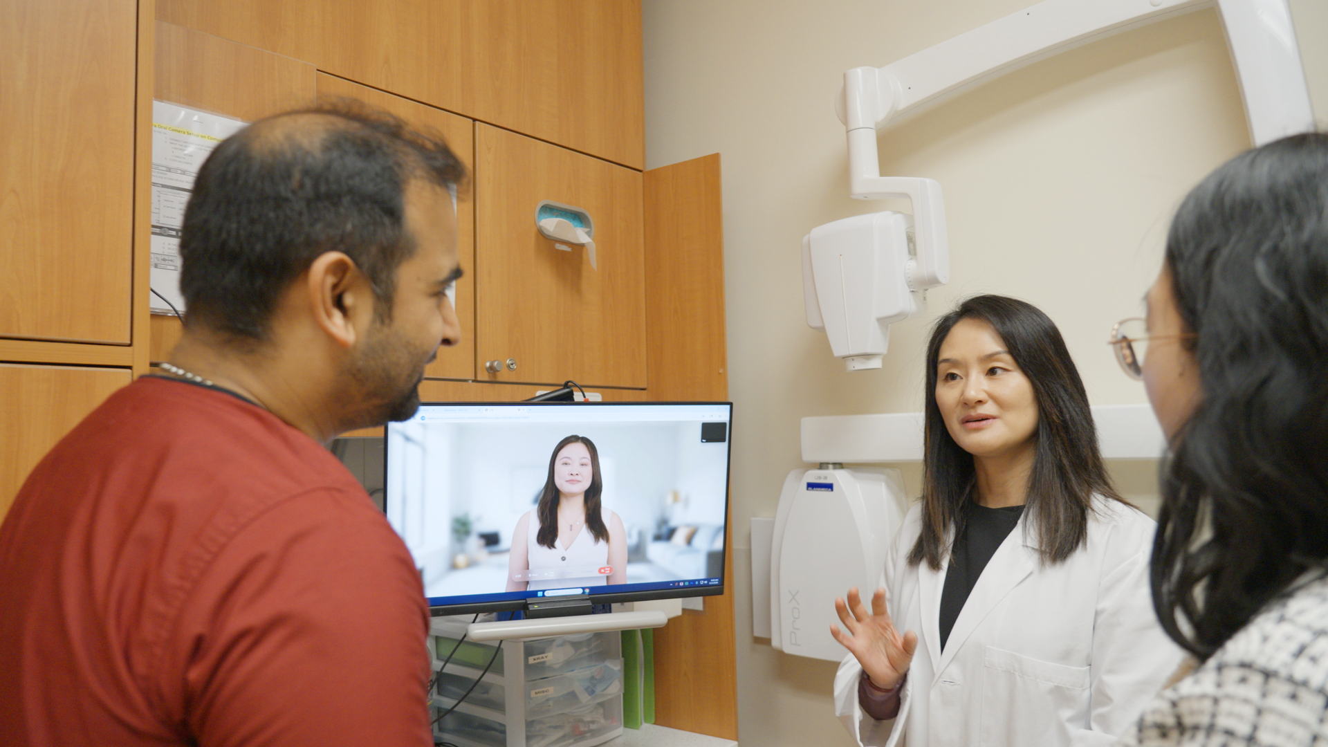 Three people talking in front of a computer screen, which displays the image of a woman.