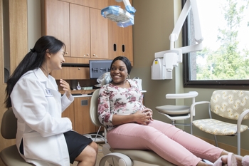 A pregnant woman with dark skin sits in a dental chair and smiles at a dentist facing her.