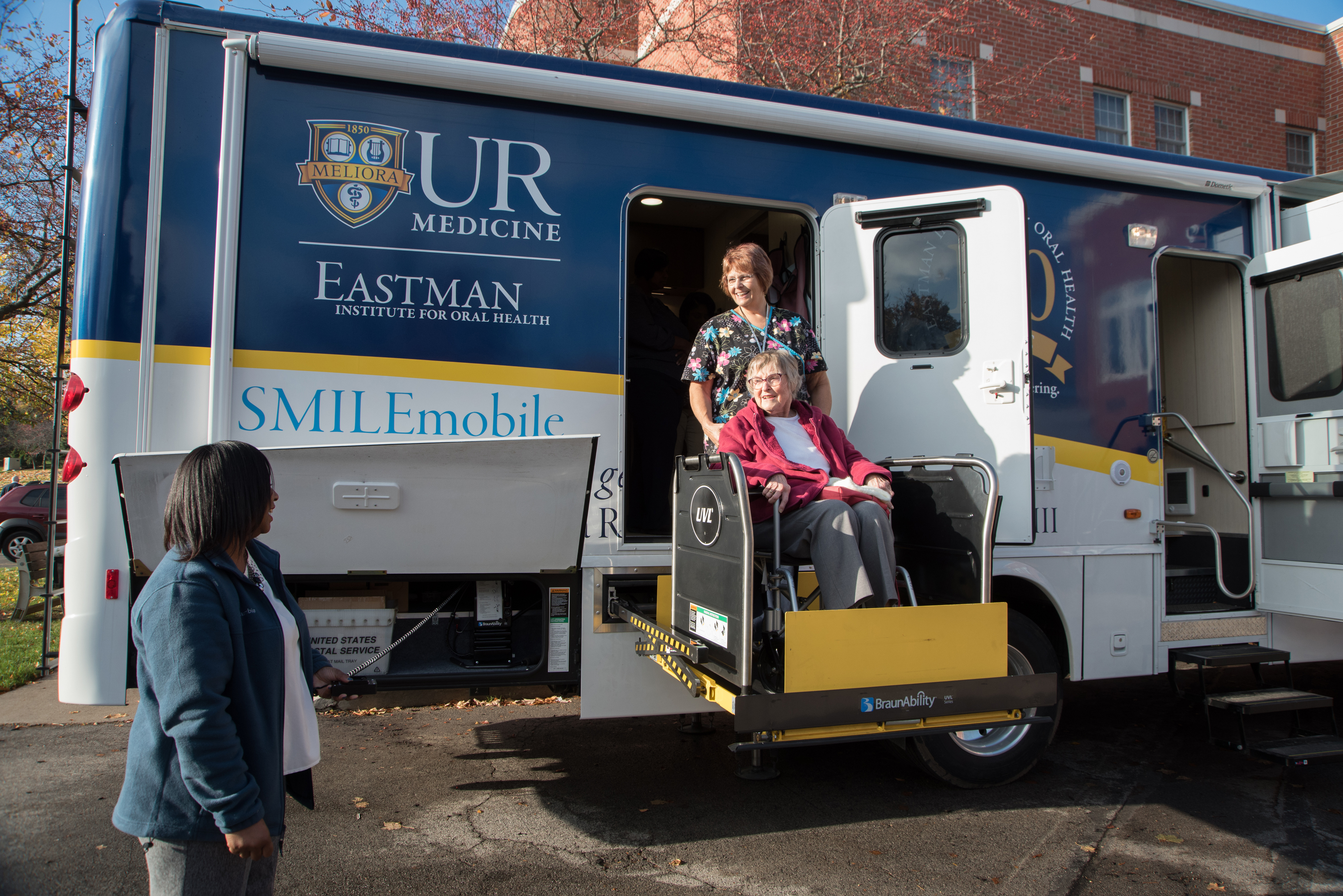 A woman in a wheelchair smiles from a wheelchair lift connected to a large vehicle resembling a camper that has "SMILEmobile" written on the side underneath the EIOH logo. A smiling woman wearing scrubs stands behind the wheelchair while another woman presses a button on the side of the vehicle.