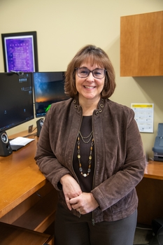 A smiling woman with short brown hair leans against a desk with a computer on it.