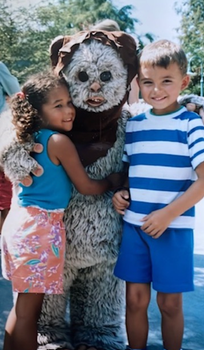 Picture of Zack and Leah as children at Disney World smiling and hugging a person dressed in a furry costume with a large, round mask and a brown hood. The setting appears to be outdoors on a sunny day with trees and other people in the background.