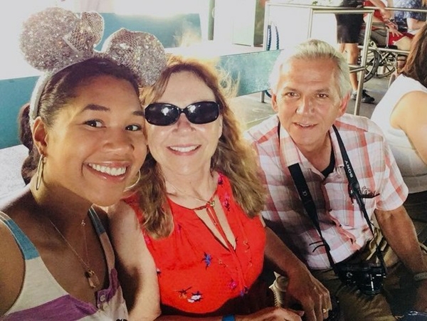 Leah with her parents, Lisa and John, sitting closely together and smiling at the camera at Disney World. One person wears sparkly mouse ears, another wears sunglasses and a red floral top, and the third has a camera around their neck, 