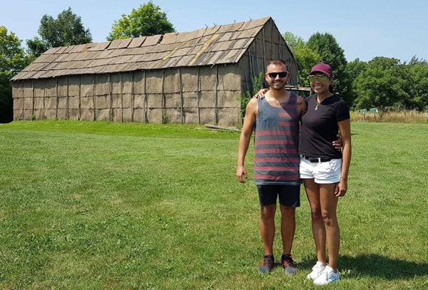 Photograph of Leah and Zack standing on green grass in front of a Ganondagan State Historic Site, surrounded by trees under a clear blue sky. Zack and Leah are casually dressed and smiling at the camera.