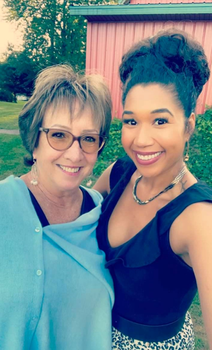 Leah and her 12 Step Sponsor Maureen posing outdoors in front of a red building and greenery. Maureen woman wears glasses and a light blue top, while Leah has a black sleeveless top and patterned skirt, both appearing happy and close.
