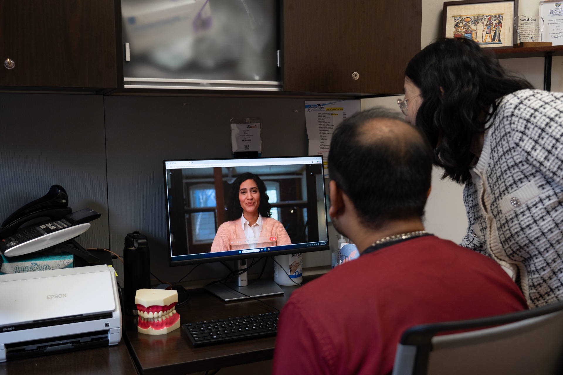A man with brown skin sits facing a computer displaying the image of a woman. A woman with black hair leans toward the computer screen from the side. 
