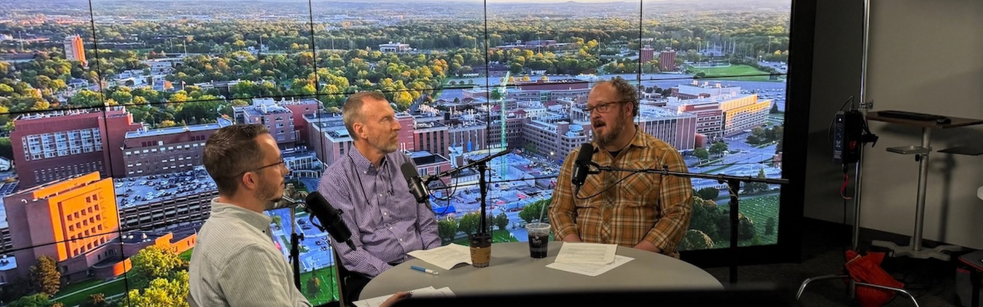 Three men sit at a table in a studio, talking and gesturing while a large window shows a wide city and harbor skyline with boats in the background.