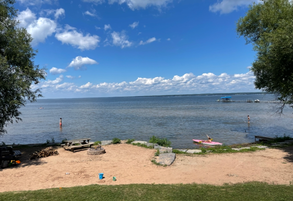 Photograph of a Oneida Lake beach scene under a partly cloudy blue sky, showing calm water with two people standing in shallow areas and one person sitting on a pink kayak near shore. Foreground includes sandy beach with picnic tables, fire pit, scattered toys, and grassy area, framed by trees on both sides.