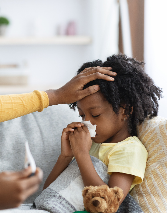 A child sitting on a couch while an adult gently wipes hair and face with a tissue as the child holds a hand near the nose, with a small toy on the cushion nearby.