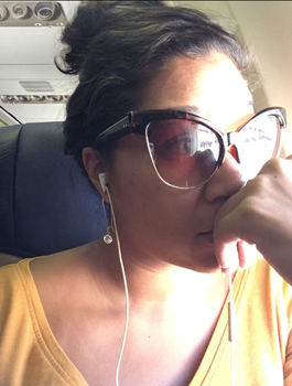Leah wearing large sunglasses and a yellow top, sitting in an airplane seat with earphones in. She appears to be looking out the window thoughtfully, with her hand resting near her mouth.