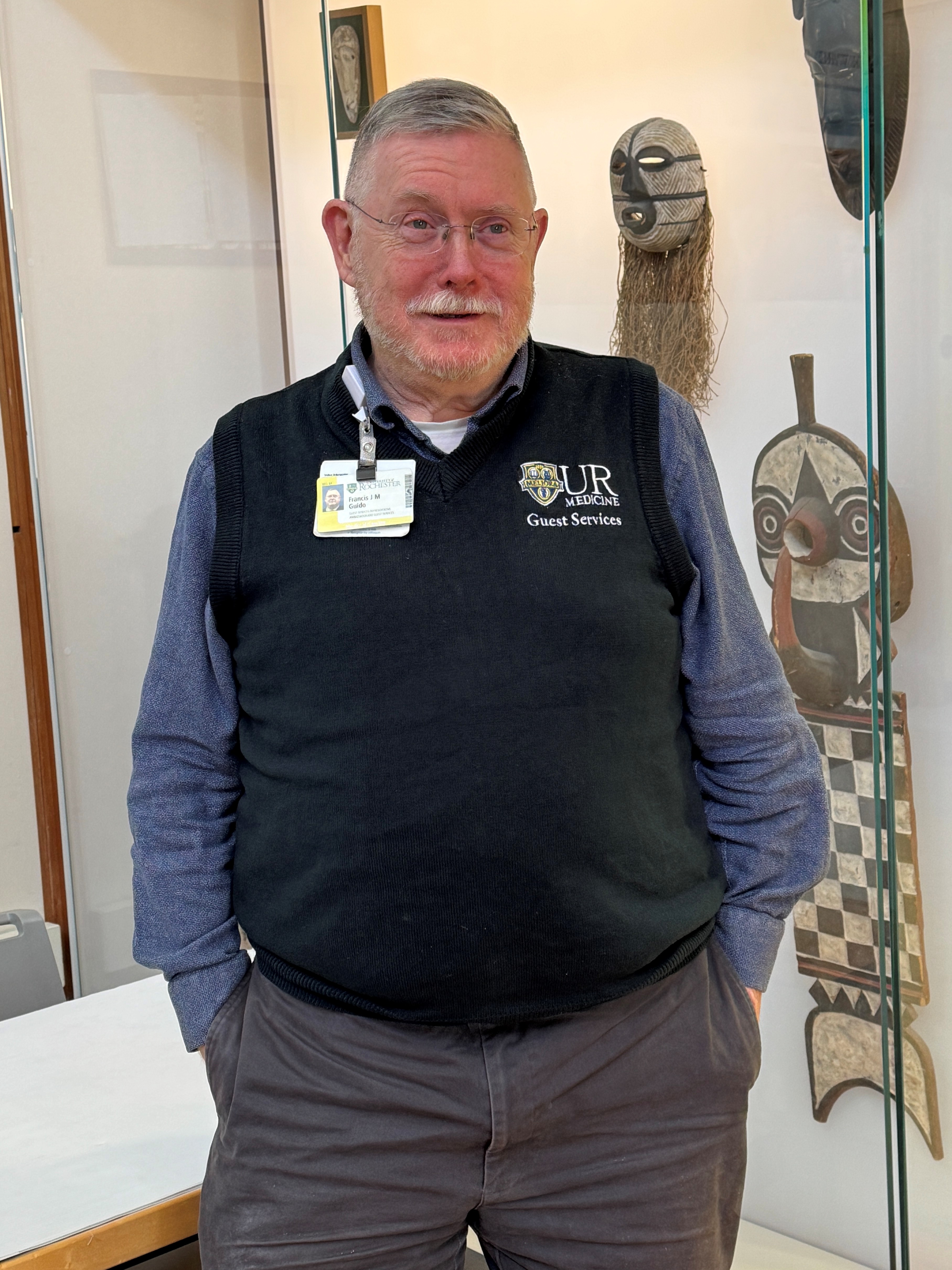 A man with short grey hair and a cropped white beard, wearing a black sweater vest with a logo for UR Medicine Guest Services.