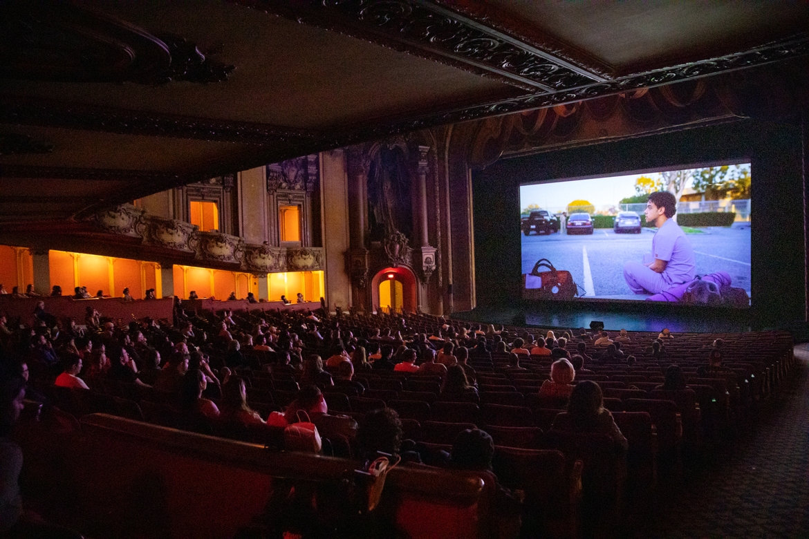 A large audience is seated in the Los Angeles Theatre watching a short film